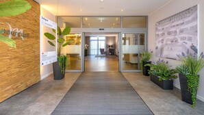A hotel lobby with a reception desk, a large poster, potted plants, and a glass door leading to a seating area. - Mercure Hotel Stuttgart Airport Messe (Stuttgart)