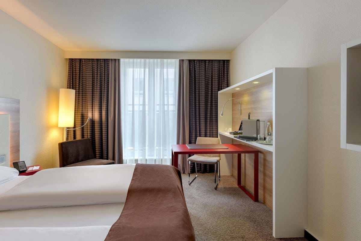 A hotel room with a bed, desk, chair, lamp, and a view of the outdoors.