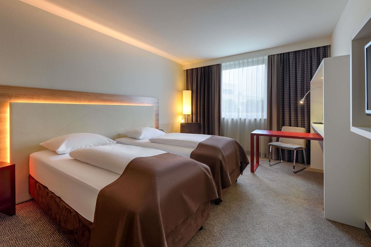 A hotel room with a bed, two bedside tables, a desk, a chair, and a large window with curtains.