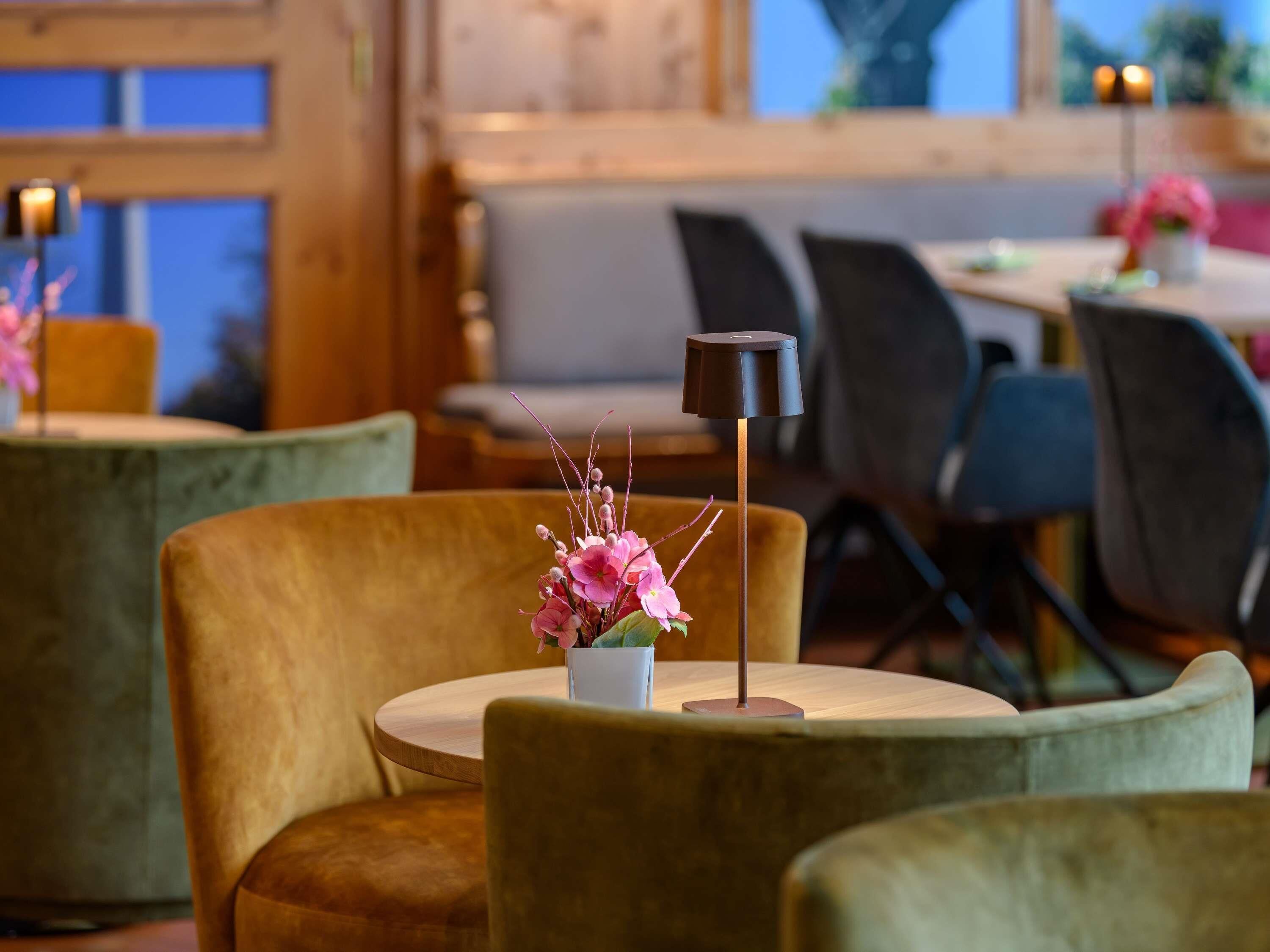 A cozy restaurant interior with round tables and plush chairs, featuring a small table with a vase of pink flowers.