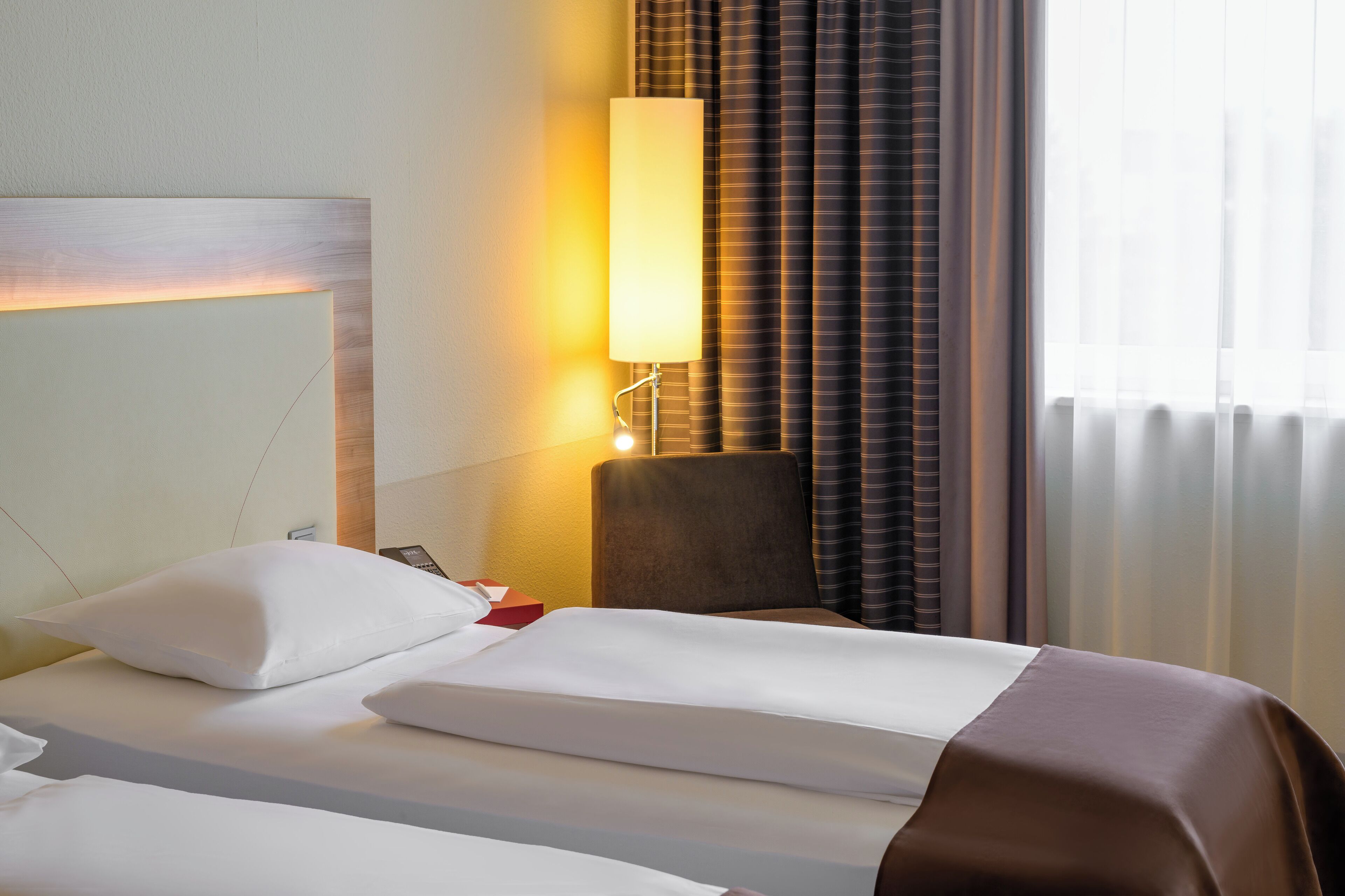 A hotel room with a neatly made bed, a bedside lamp, a chair, and a window with curtains.
