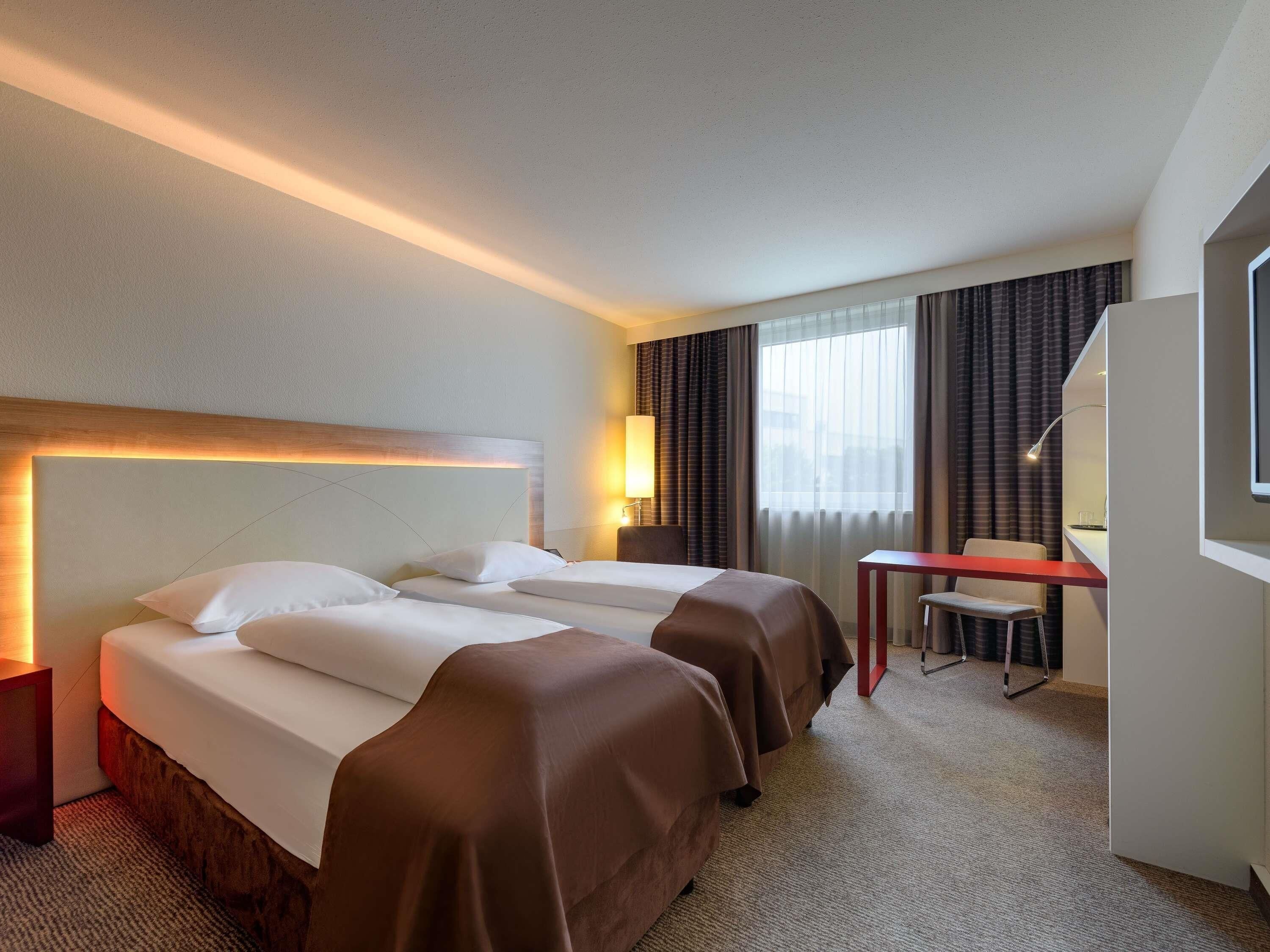 Une chambre d’hôtel comprenant un lit, deux tables de chevet, un bureau, une chaise et une grande fenêtre avec des rideaux.