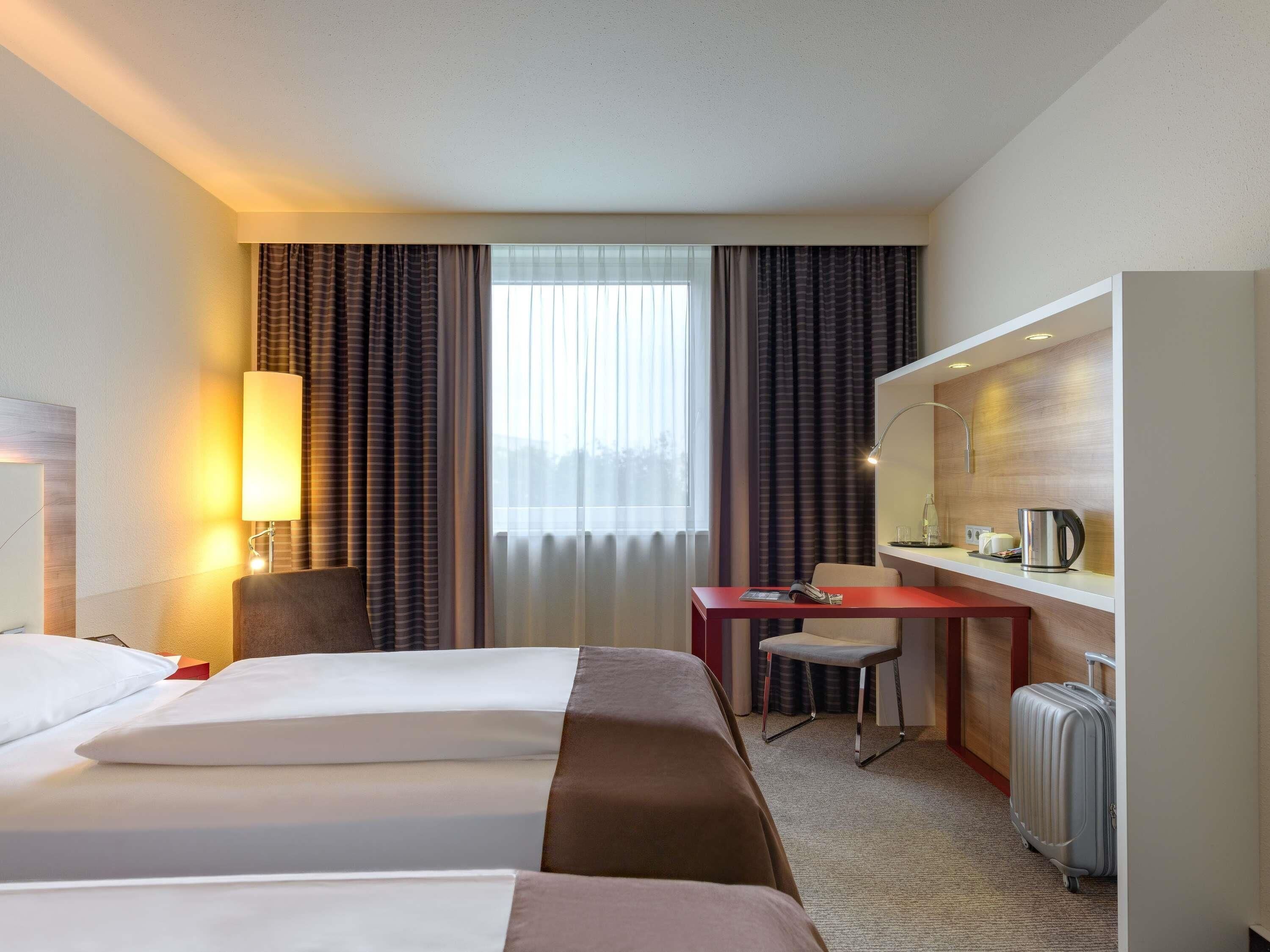 A hotel room with two beds, a desk, a chair, a small table with a kettle, and a suitcase.