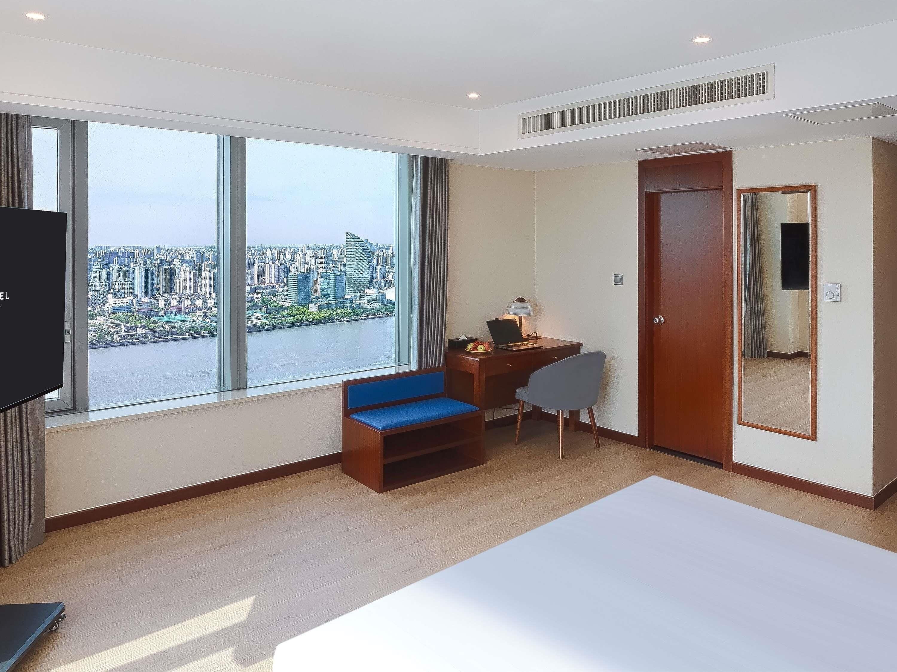A hotel room with a large window, a desk with a chair, a bed, and a wardrobe.