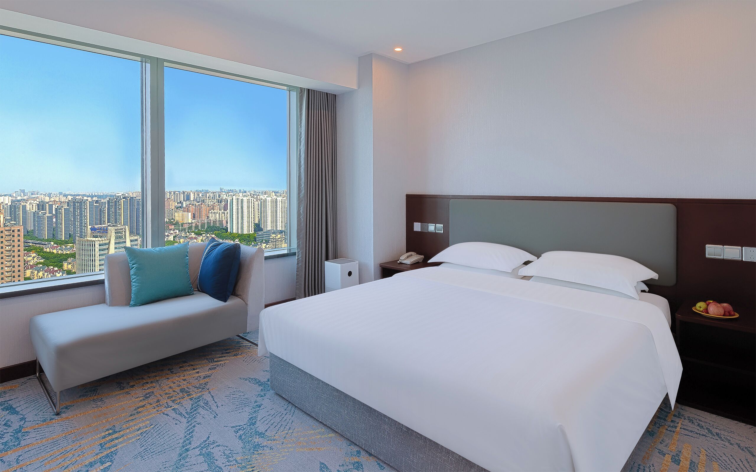 A hotel room with a large bed, a sofa with pillows, a bedside table with a phone, and a view of a cityscape through large windows.