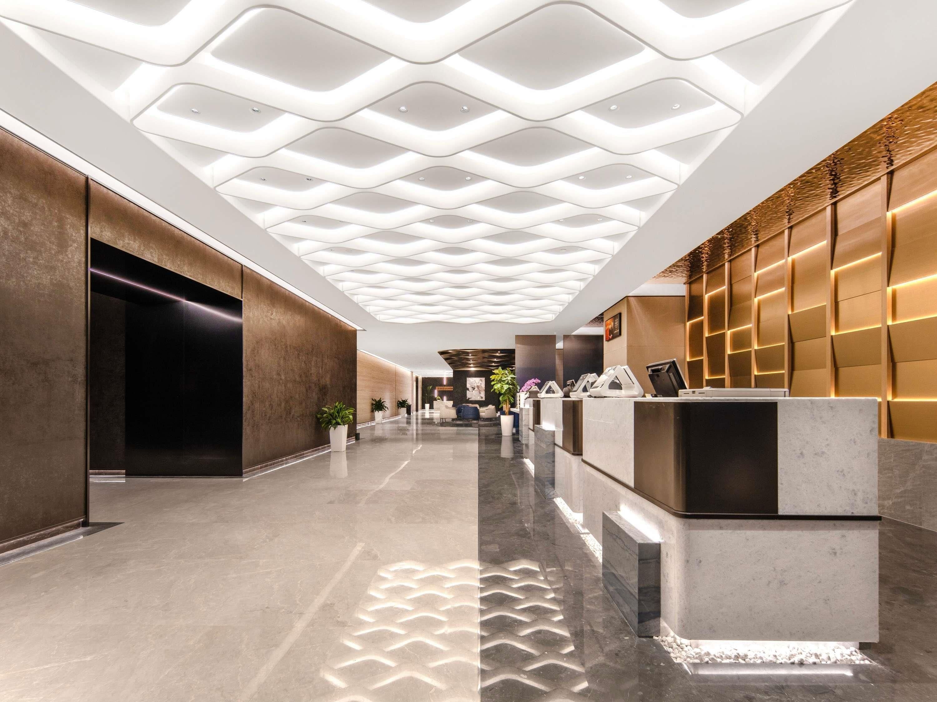 A modern lobby with a geometric ceiling, a reception desk, and a plant.