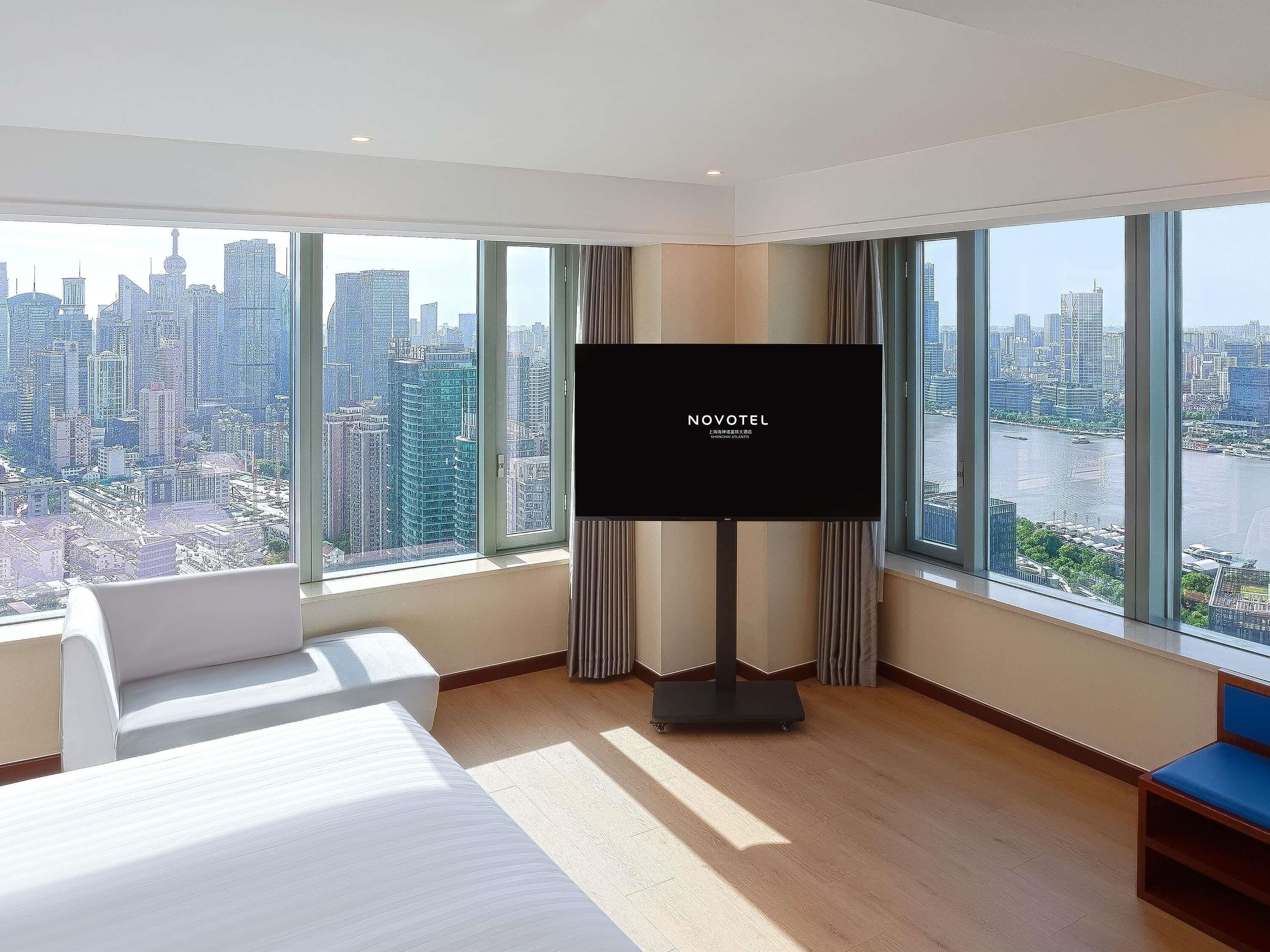 Hotelkamer met een groot televisiescherm, een bed, een bank en uitzicht op een stadsgezicht door grote ramen.
