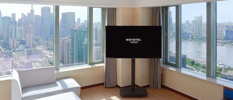 Hotelkamer met een groot televisiescherm, een bed, een bank en uitzicht op een stadsgezicht door grote ramen.