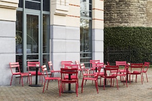 Outdoor red chairs and tables arranged in front of a building with large windows and a brick wall.