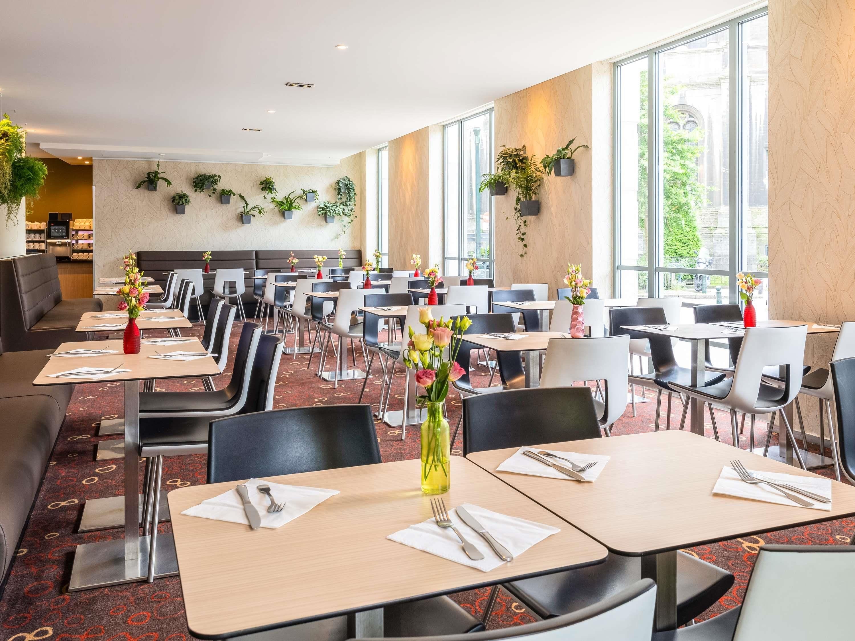 A spacious dining area with multiple tables and chairs, large windows, and potted plants on the walls.