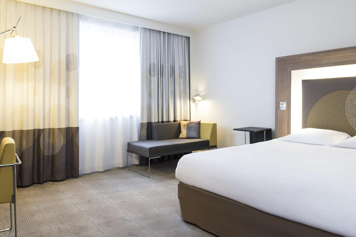 Une chambre d’hôtel avec un lit, un bureau, une chaise, une lampe et une fenêtre avec des rideaux.