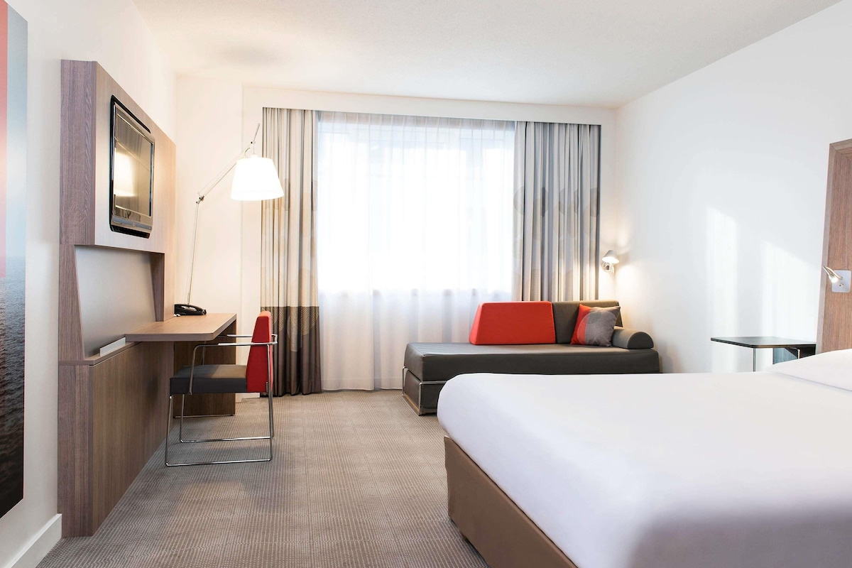 Une chambre d’hôtel équipée d’un lit, d’un bureau, d’une chaise, d’un canapé, d’une télévision et d’une fenêtre avec des rideaux.