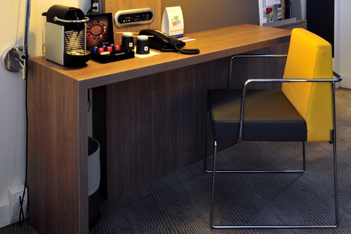 A hotel room with a wooden desk, a television mounted on the wall, a desk lamp, a chair, and a small trash bin.