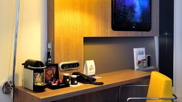 Een hotelkamer met een houten bureau, een aan de muur bevestigde televisie, een bureaulamp, een stoel en een kleine prullenbak.