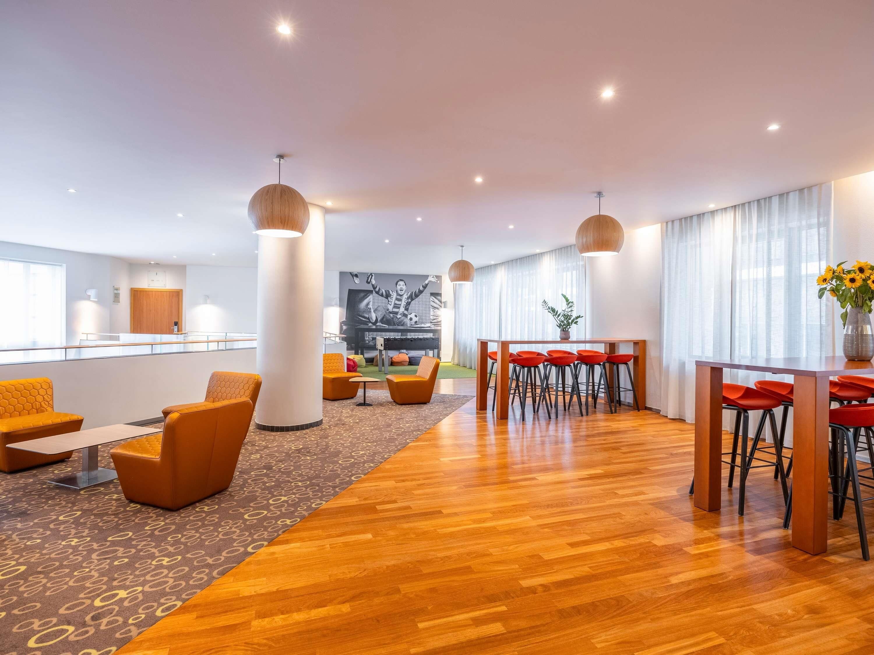 Un hall d’hôtel moderne avec un plancher en bois, des sièges orange, un piano et une grande fenêtre avec des rideaux.