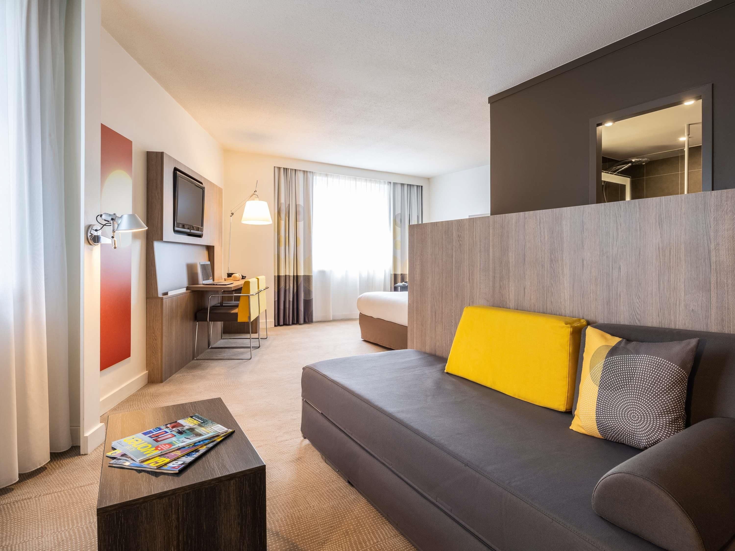 Une chambre d’hôtel moderne équipée d’un canapé, d’une petite table, d’un bureau avec une chaise, d’une télévision et d’une grande fenêtre avec des rideaux.
