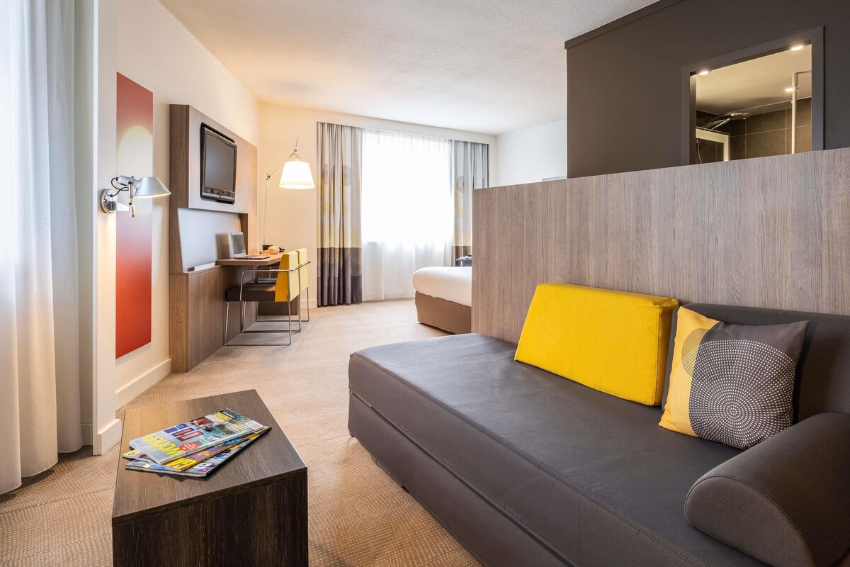 Una camera d'albergo moderna con un divano, un tavolino, una scrivania con una sedia, una TV e un'ampia finestra con tende.
