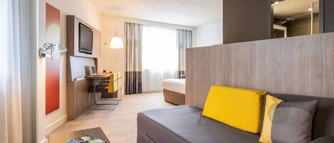 A modern hotel room with a sofa, a small table, a desk with a chair, a TV, and a large window with curtains.