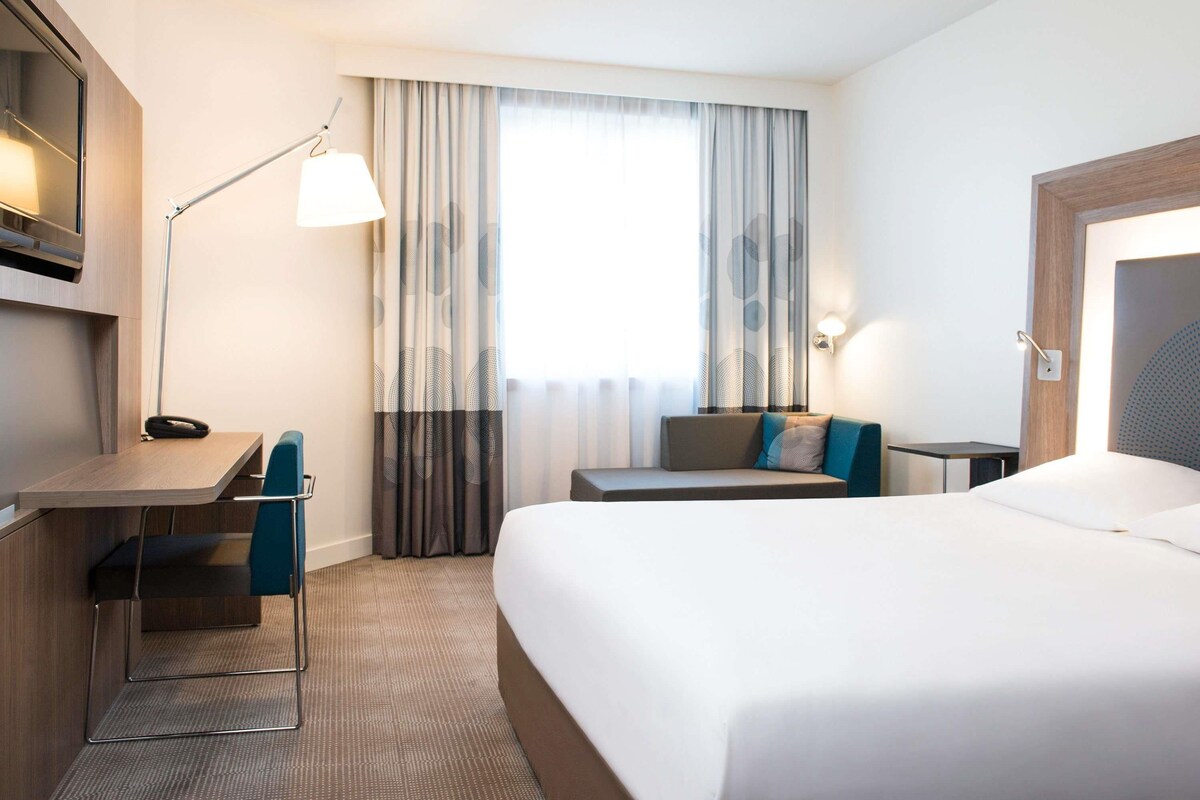 A hotel room with a bed, desk, chair, lamp, and curtains.