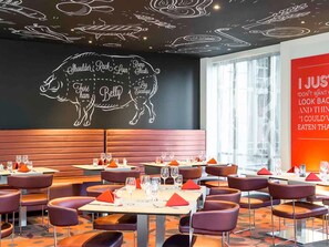 Interior de restaurante con una pared de pizarra mostrando los artículos del menú y un gran letrero rojo con texto. Las mesas están dispuestas con vasos y servilletas.