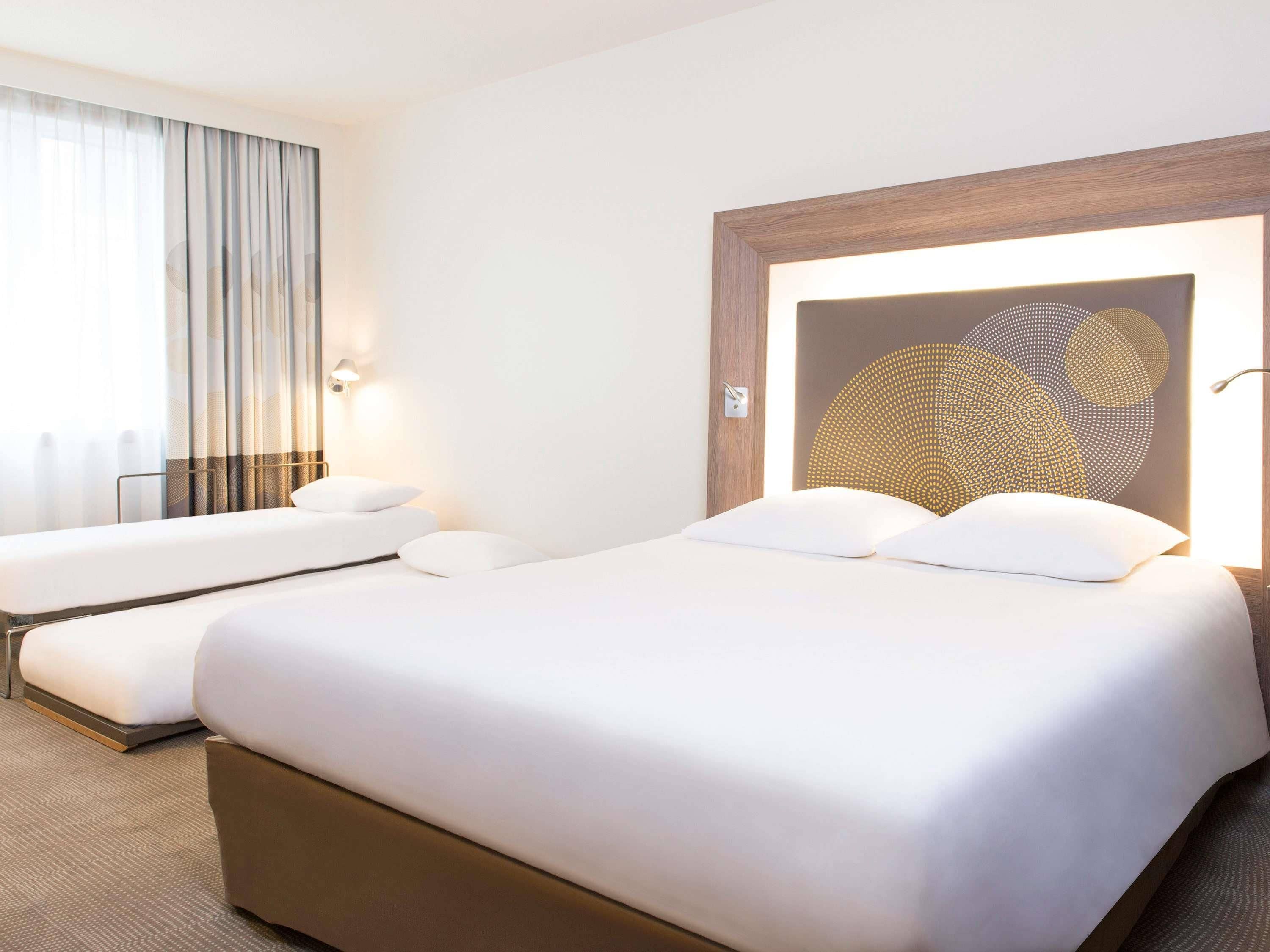 A hotel room with two beds, a large window with curtains, and a wall-mounted light fixture.