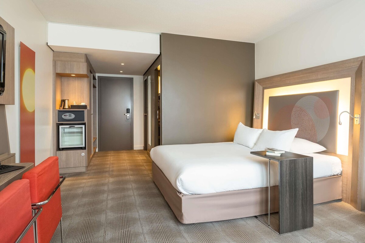 Una camera d'albergo con un letto, una scrivania, una poltrona rossa, un minibar e una TV a parete.