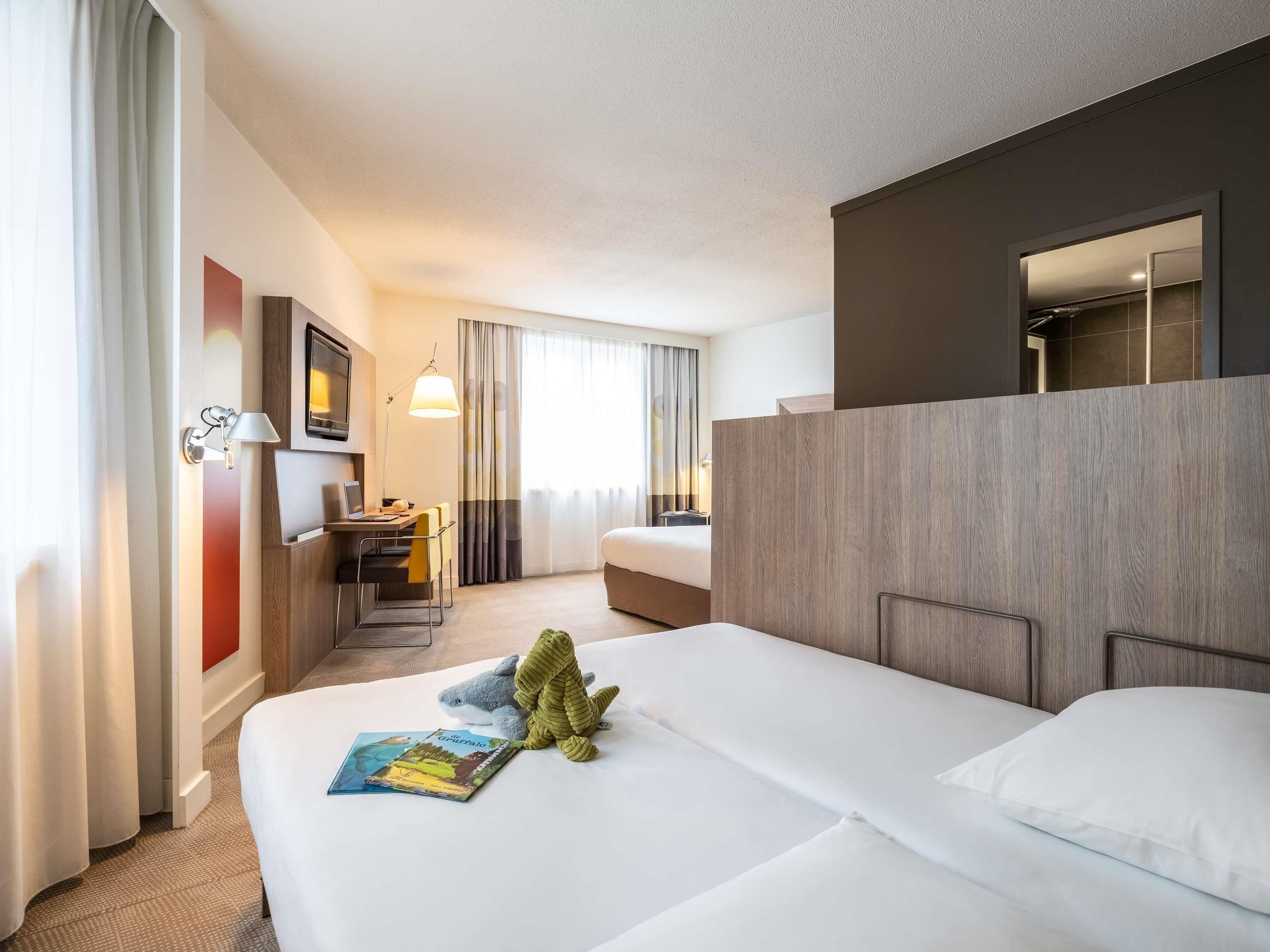 Quarto de hotel com cama, escrivaninha com cadeira, televisão e janela com cortinas.