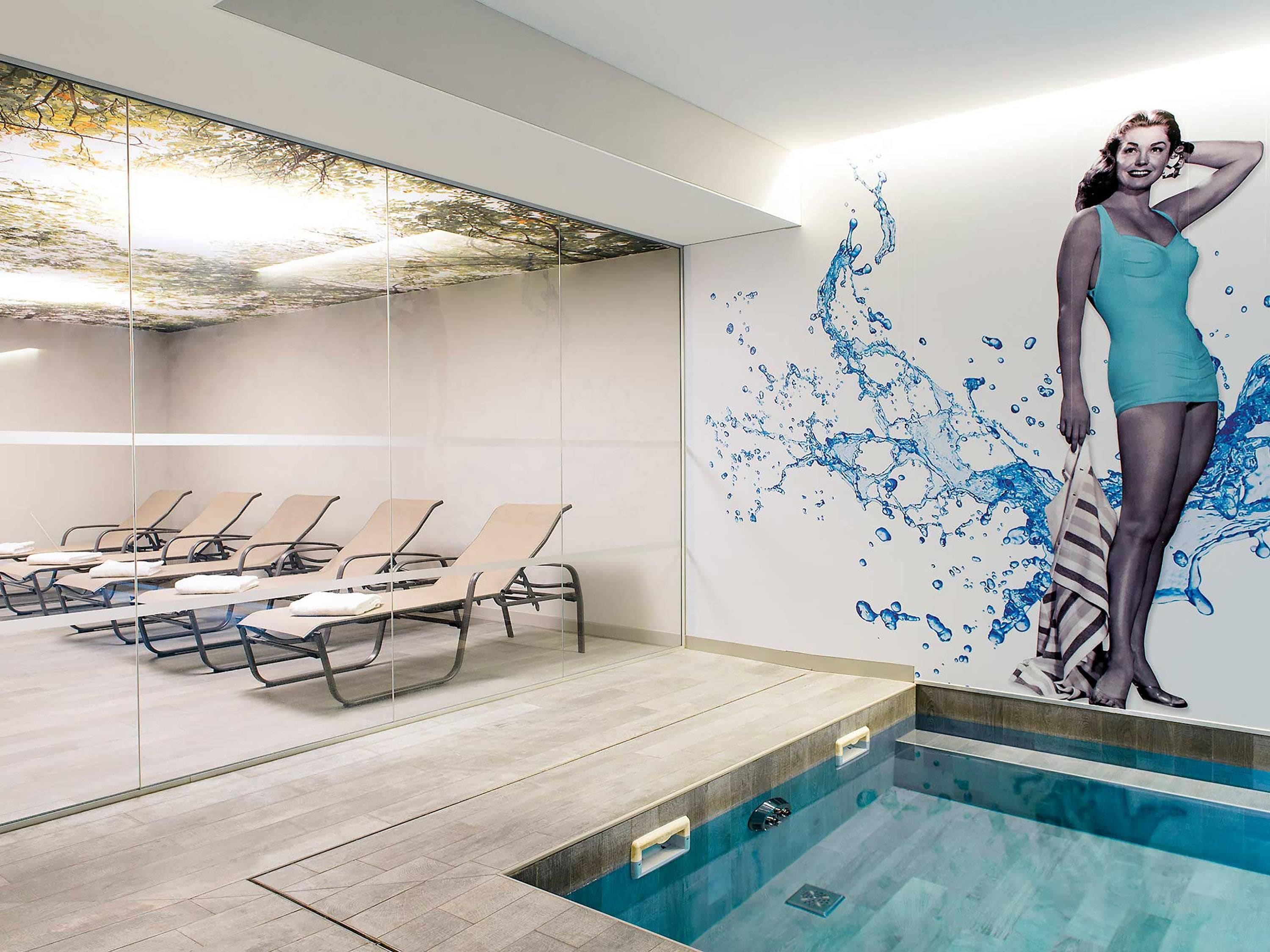 Un espace piscine intérieure moderne, doté de chaises longues et d’une grande fresque représentant une femme en maillot de bain.