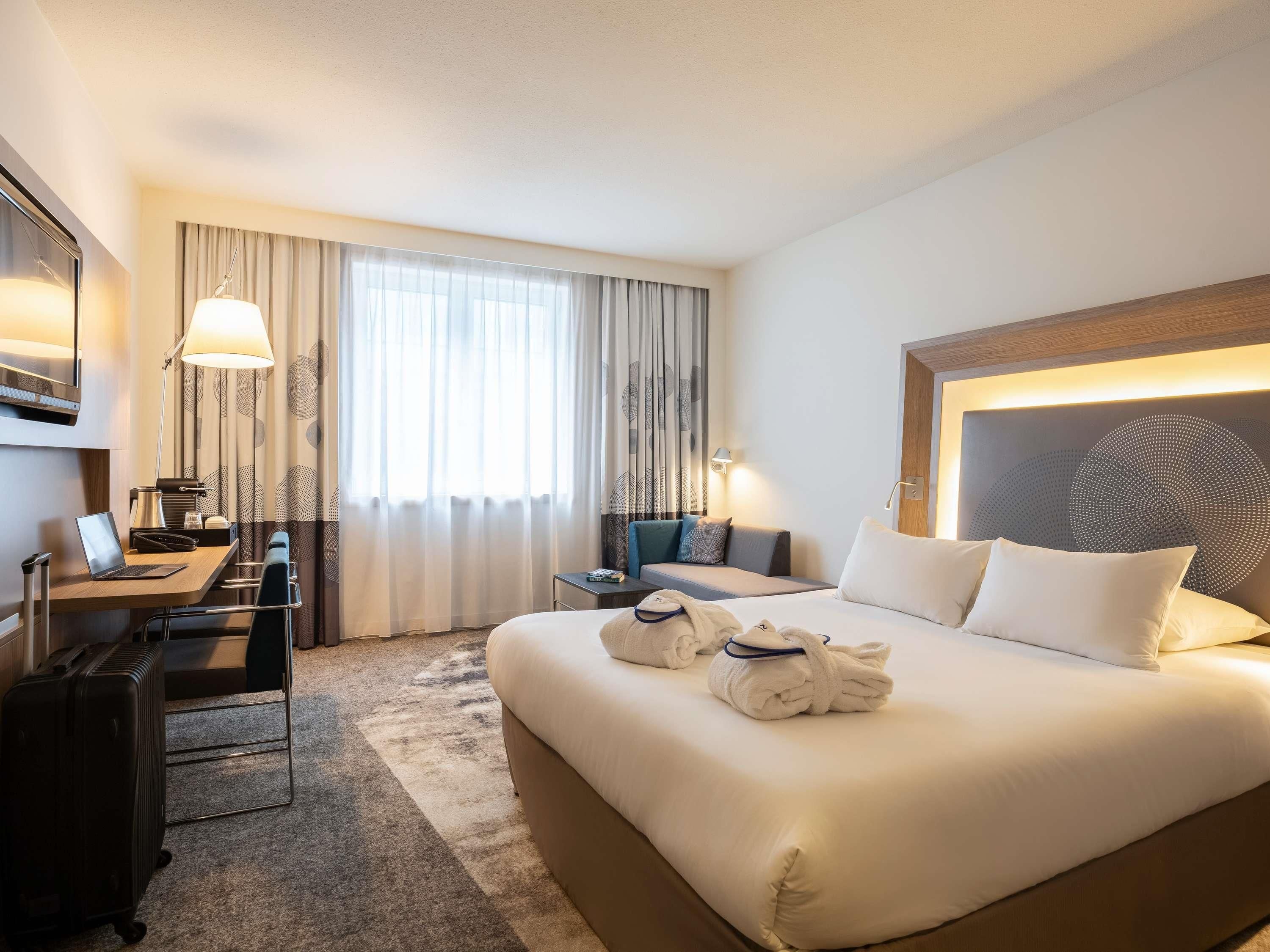 A hotel room with a large bed, a desk with a laptop, a chair, a suitcase, and a window with curtains.