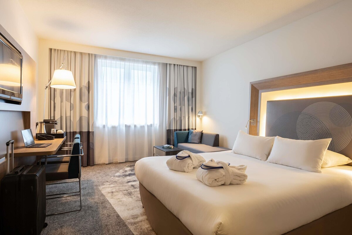 A hotel room with a large bed, a desk with a laptop, a chair, a suitcase, and a window with curtains.