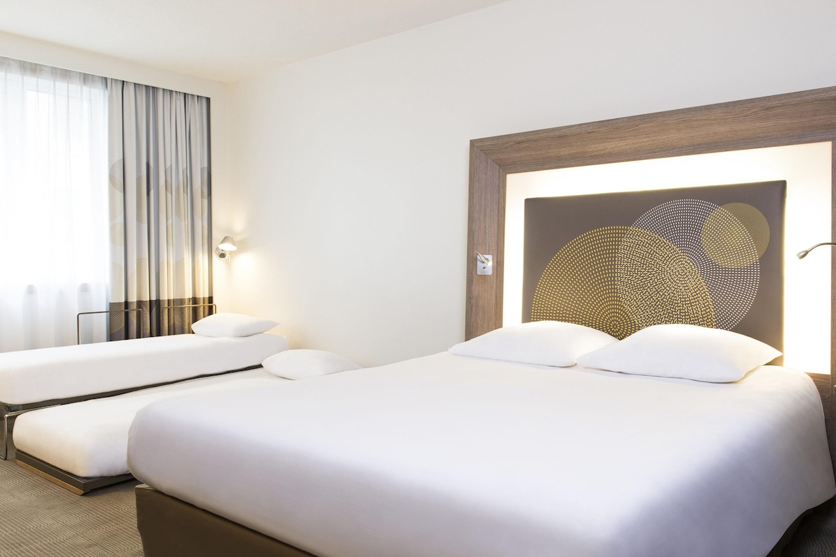 A hotel room with a large bed, a smaller bed, a wall-mounted lamp, and a window with curtains.