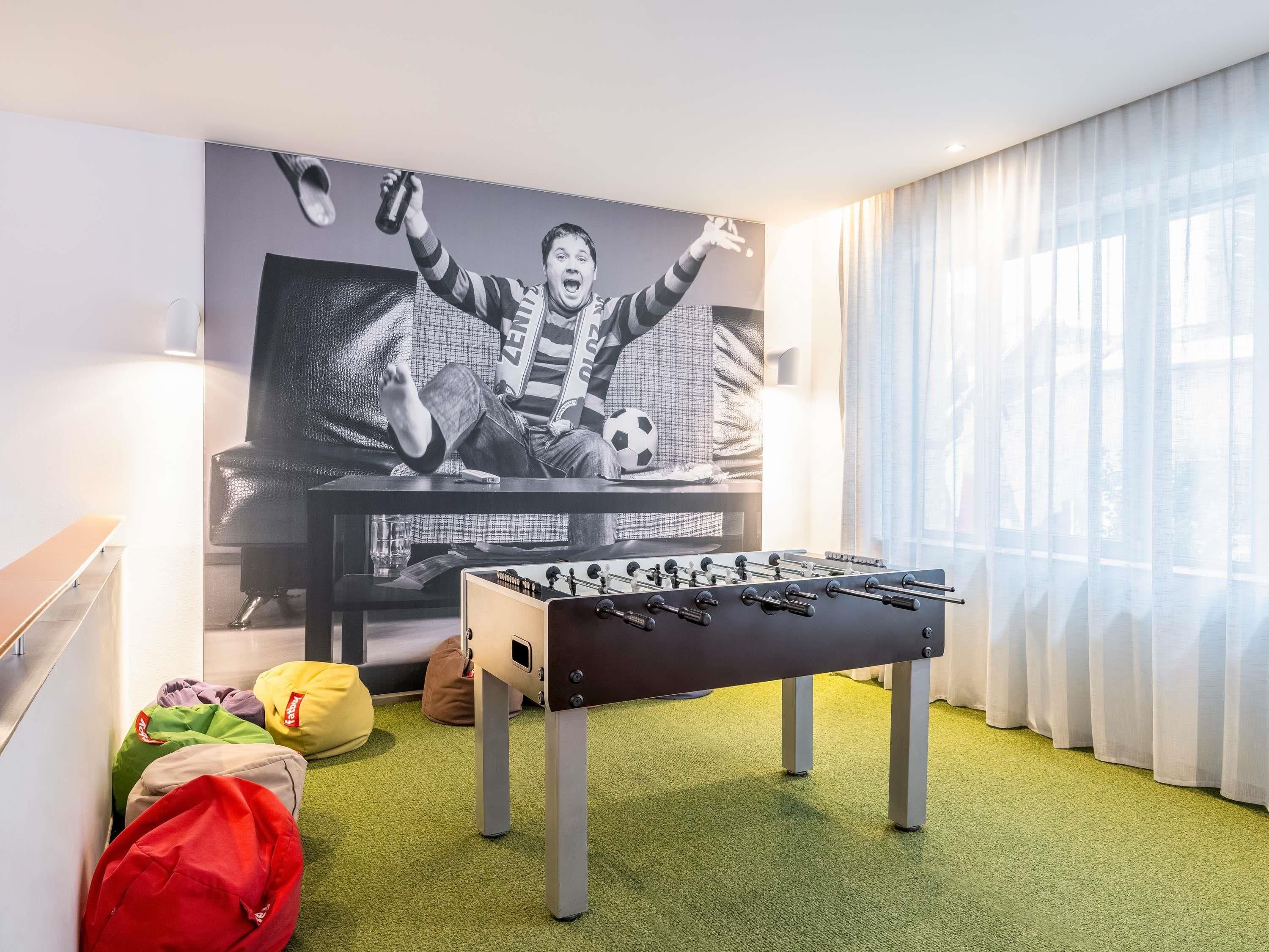 Een kamer met een tafelvoetbaltafel, een bank en een grote televisie waarop een voetballer te zien is.