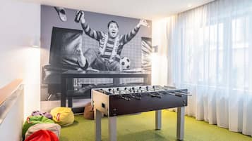 Een kamer met een tafelvoetbaltafel, een bank en een grote televisie waarop een voetballer te zien is.