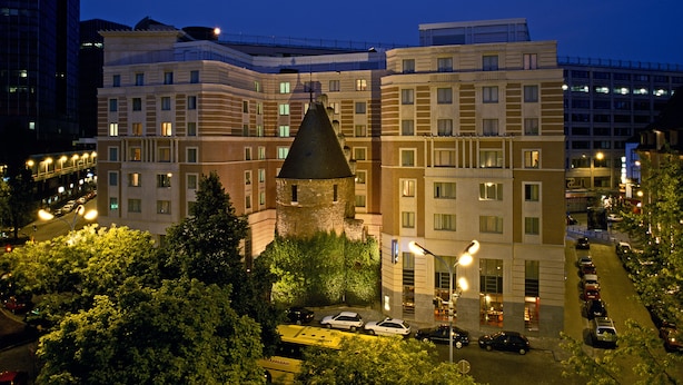 Un hôtel à plusieurs étages, doté d’une tour centrale, entouré d’arbres et de voitures garées.
