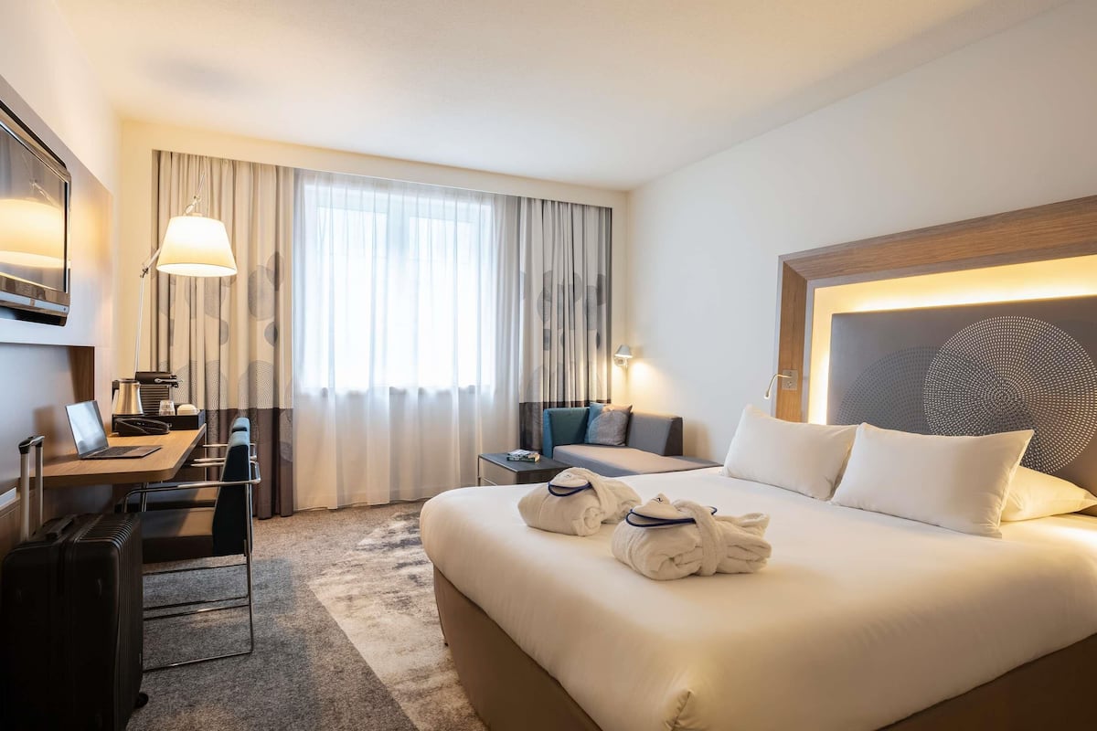 Une chambre d’hôtel avec un grand lit, un bureau avec un ordinateur portable, une chaise, une valise et une fenêtre avec des rideaux.