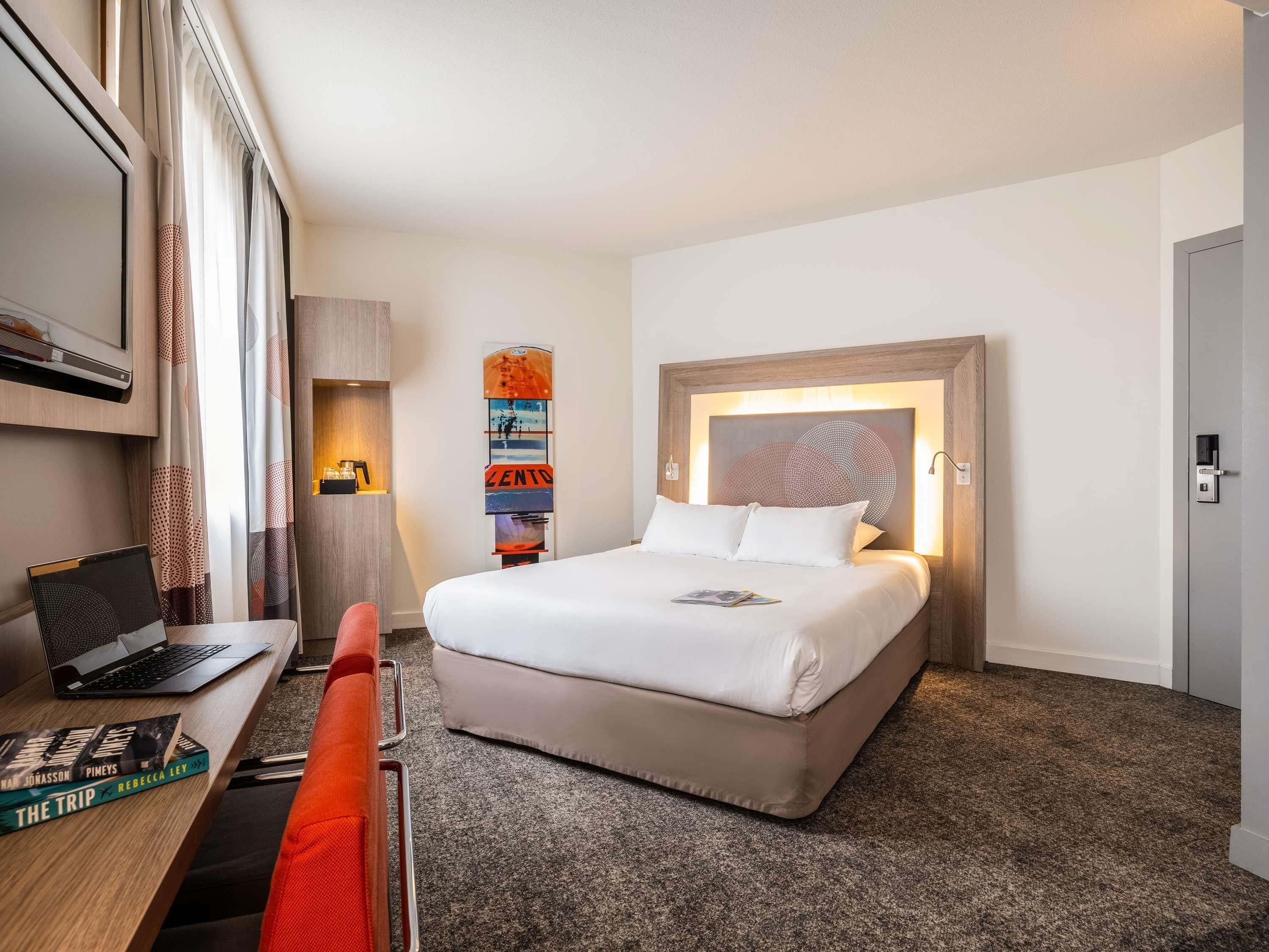 A hotel room with a bed, a desk with a laptop, a bookshelf, and a wall-mounted artwork.