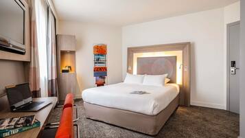 A hotel room with a bed, a desk with a laptop, a bookshelf, and a wall-mounted artwork.