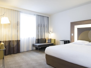 Une chambre d’hôtel avec un lit, un bureau, une chaise, une lampe et une fenêtre avec des rideaux.
