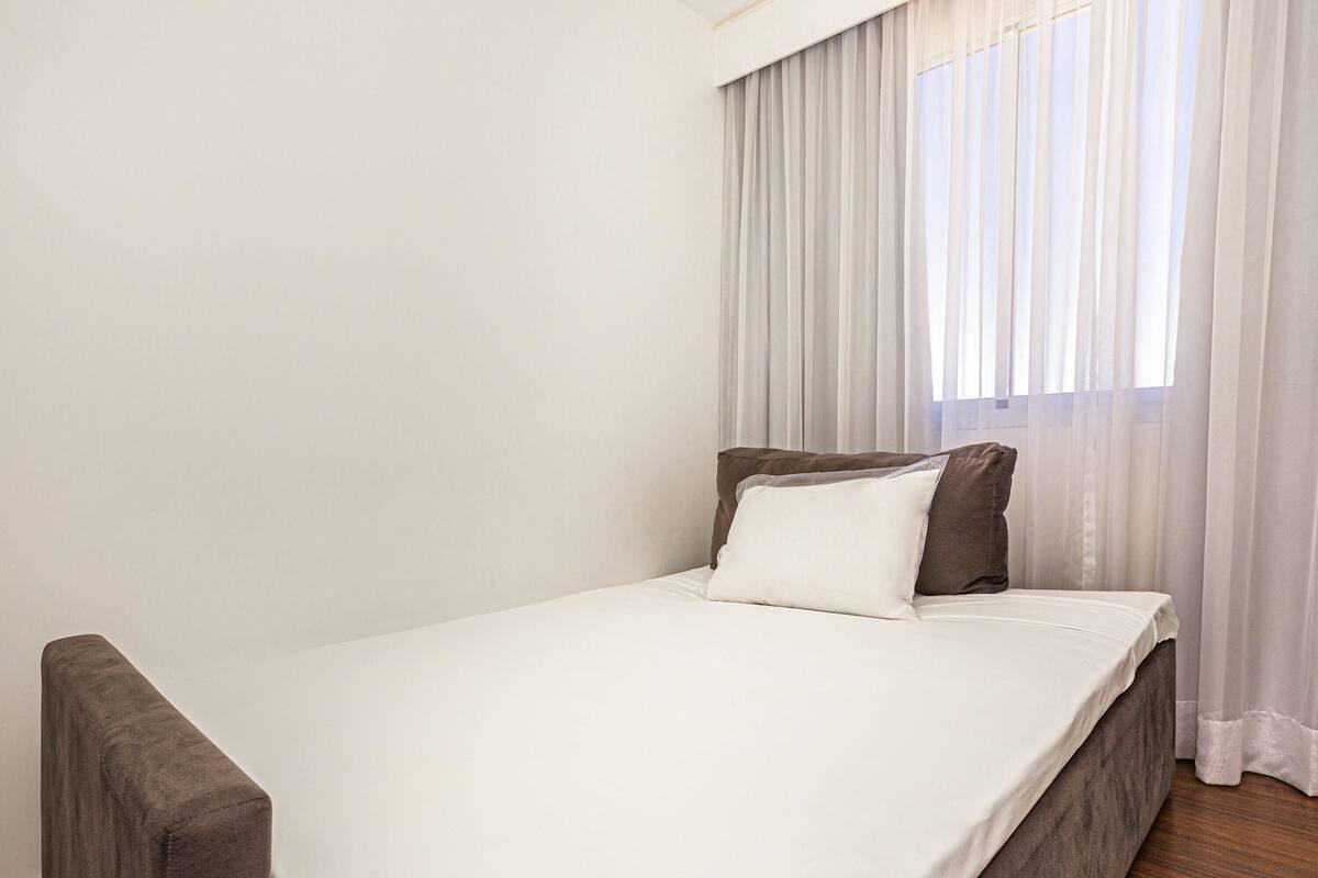 A simple, unadorned room with a single bed, a small bench, and a window with sheer curtains.