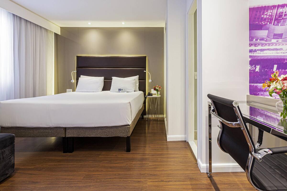 A hotel room with a large bed, a desk, a chair, and a wall-mounted TV.
