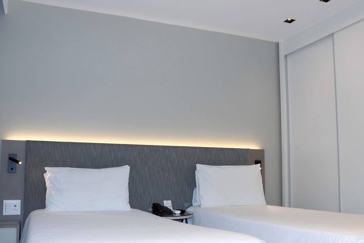 A hotel room with two beds, a bedside table with a phone, and a wall-mounted light.