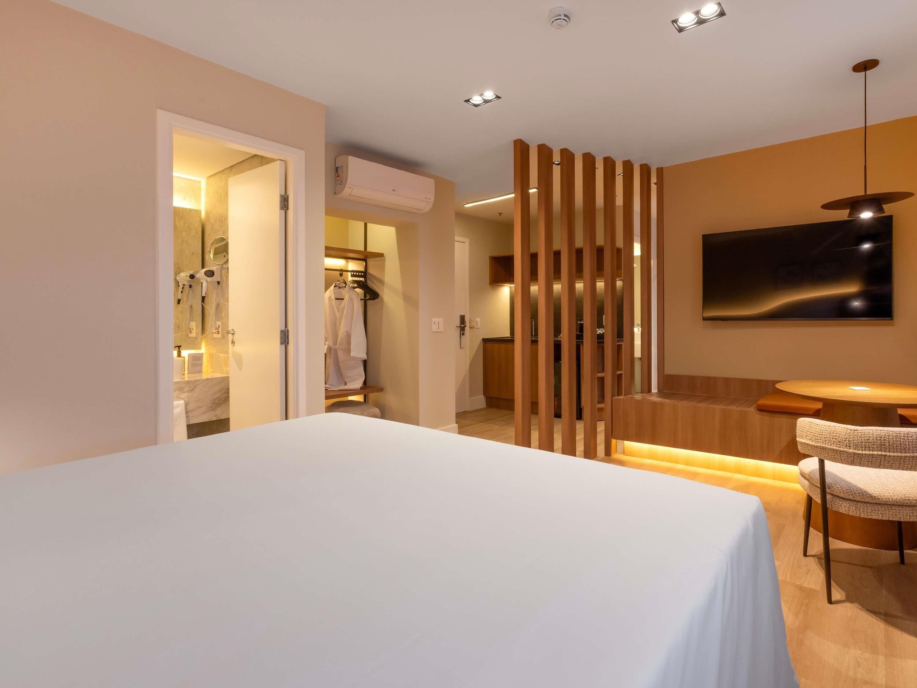 A hotel room with a bed, a seating area with a table and chairs, a shower area with a towel rack, and a mounted television.