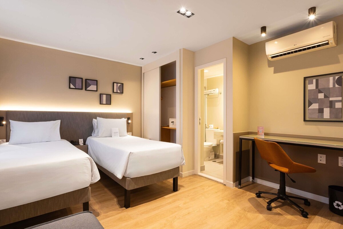 A hotel room with two beds, a desk, a chair, and an air conditioning unit.