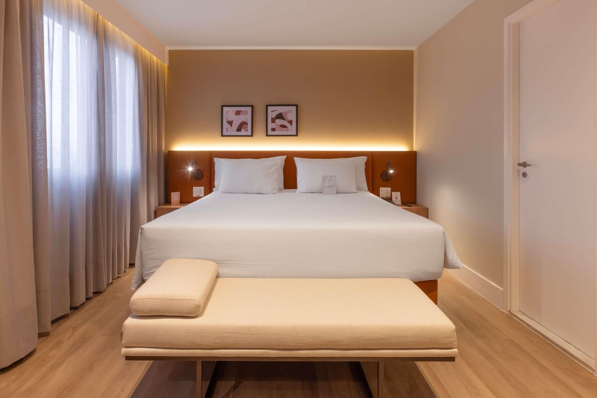 A hotel room with a large bed, bedside tables, a bench, and curtains.