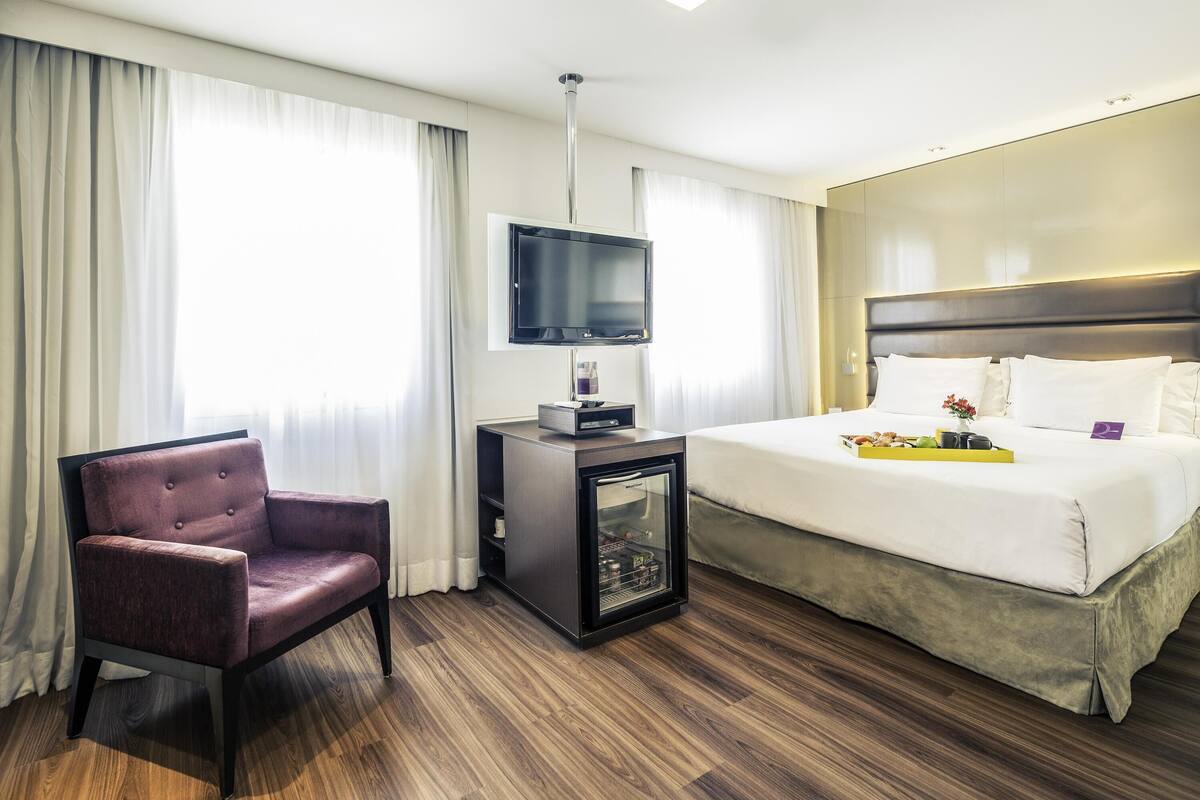 A hotel room with a bed, a TV, a chair, and a small table with a tray.