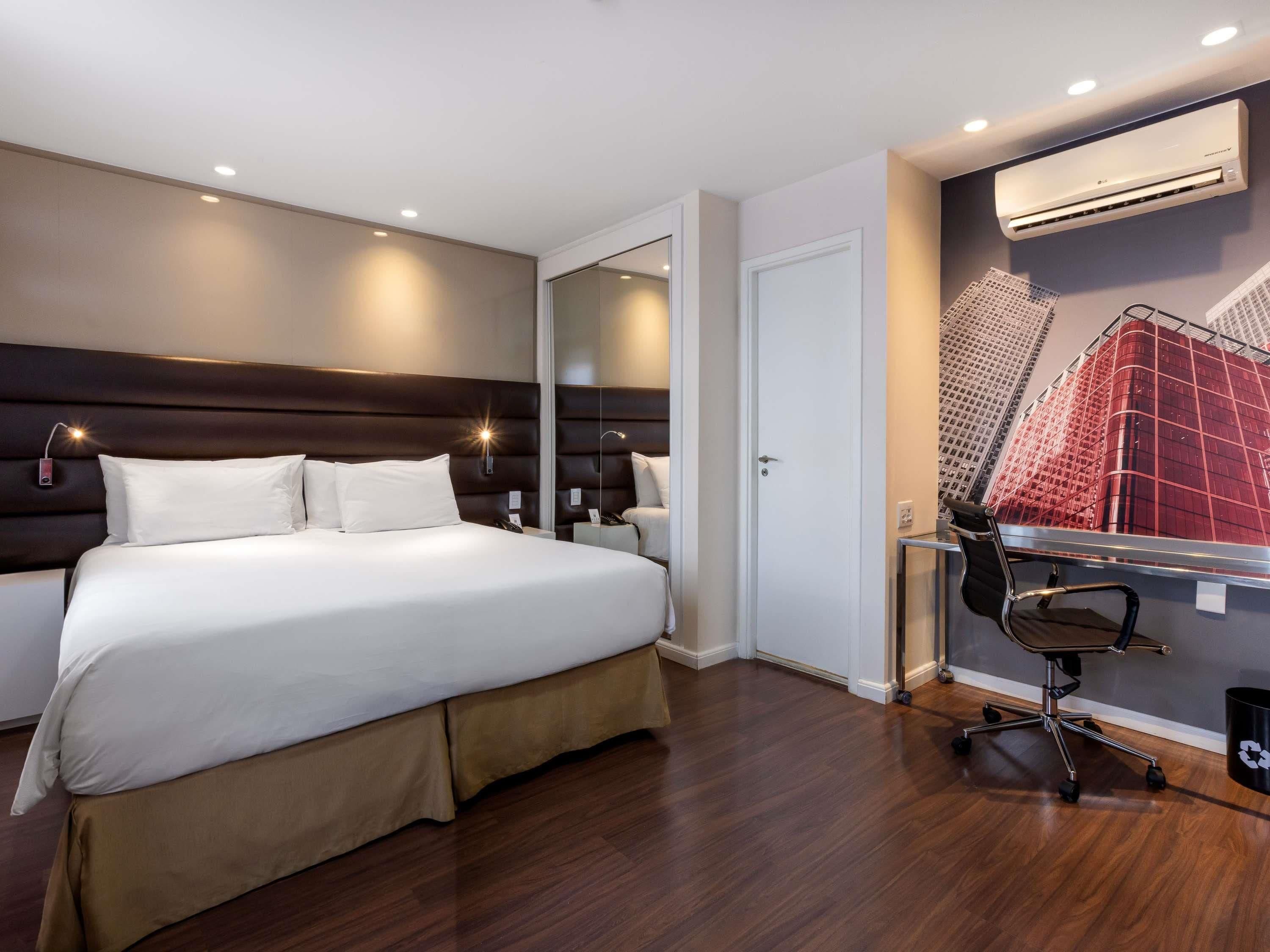 Una habitación de hotel con una cama grande, un escritorio con una silla, un equipo de aire acondicionado y un espejo.