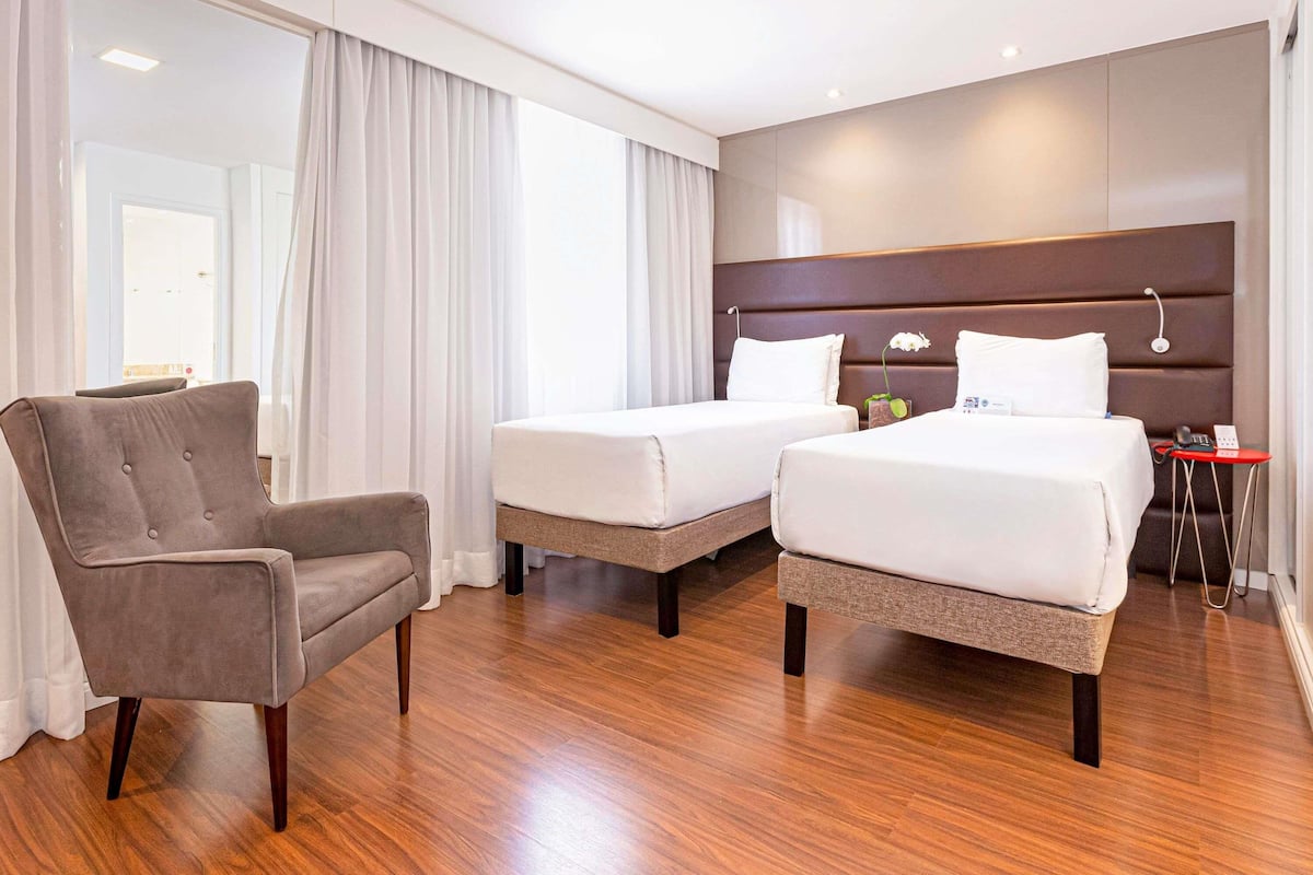 A hotel room with two beds, a chair, a bedside table with a phone, and a window with curtains.