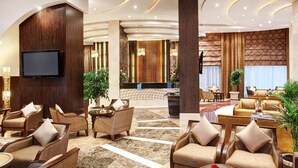 A spacious lobby with multiple seating areas, a central reception desk, and a large flat-screen TV mounted on the wall.