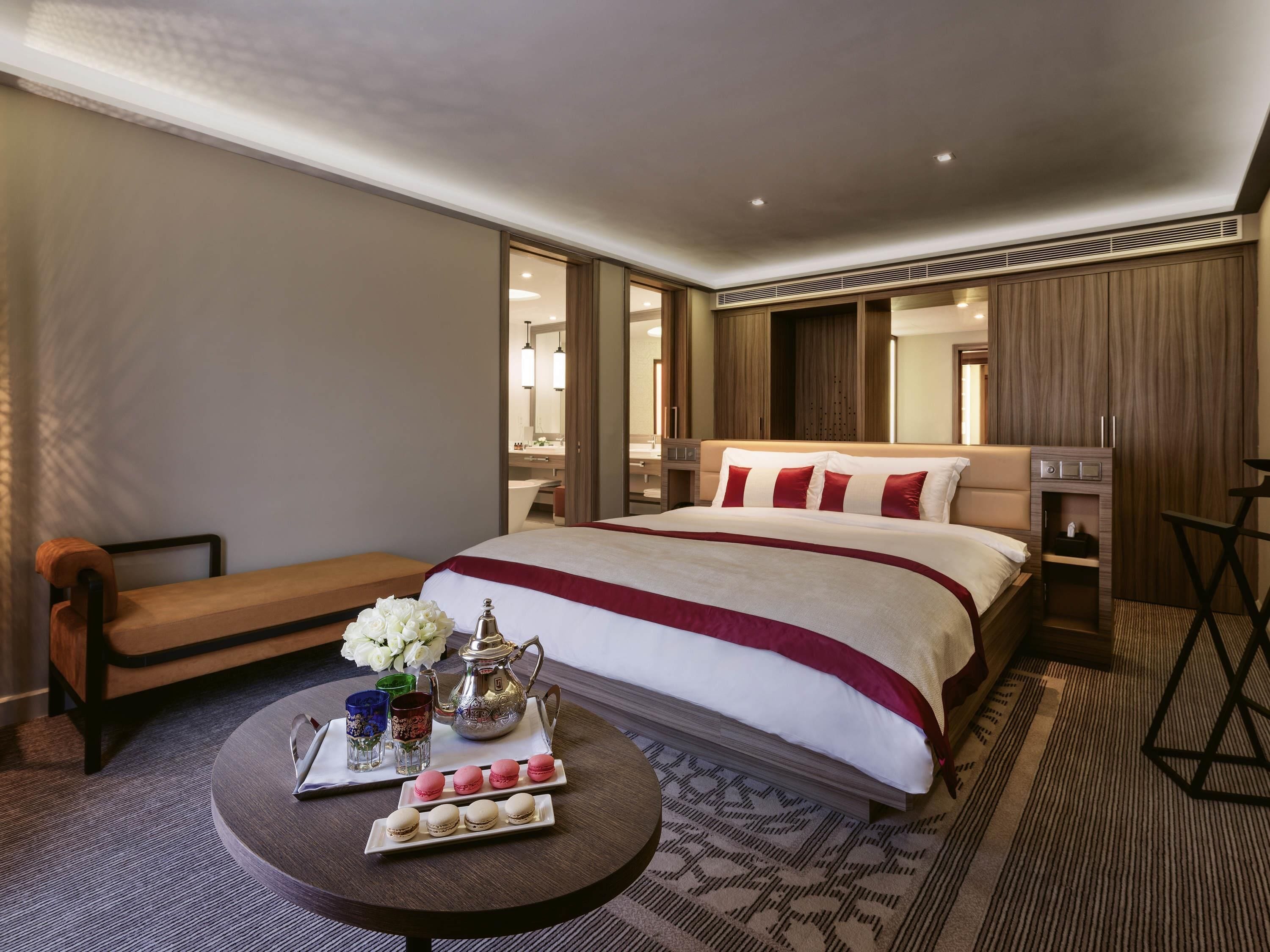 Habitación de hotel con una cama grande, mesita de noche, una pequeña mesa redonda con aperitivos, un espejo y un armario.