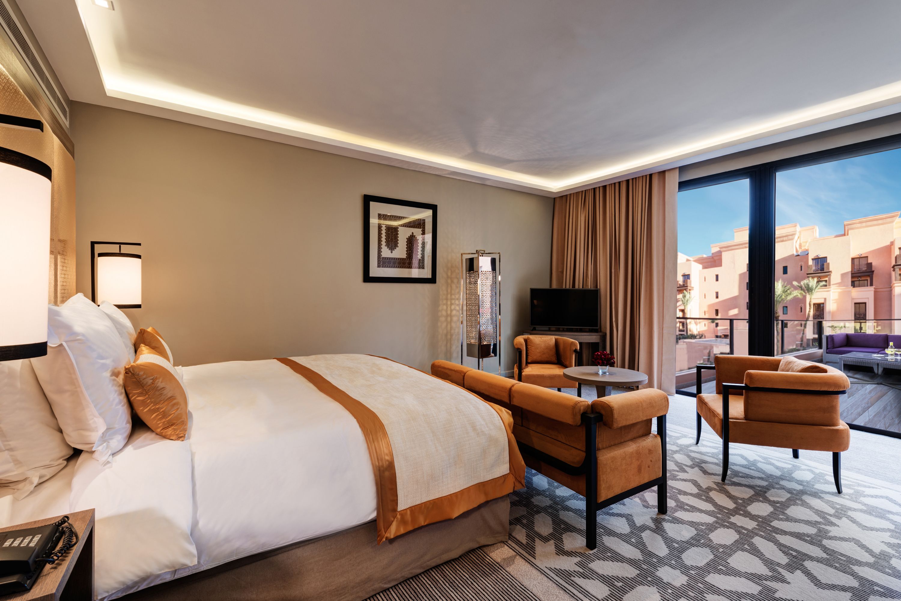 Ein Hotelzimmer mit einem großen Bett, zwei Sesseln, einem kleinen Tisch, einem Fernseher und einem großen Fenster mit Blick auf Gebäude.